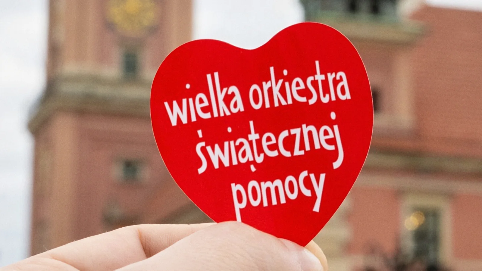 Czerwone serduszko Wielkiej Orkiestry Świątecznej Pomocy trzymane w dłoni na tle Zamku Królewskiego w Warszawie